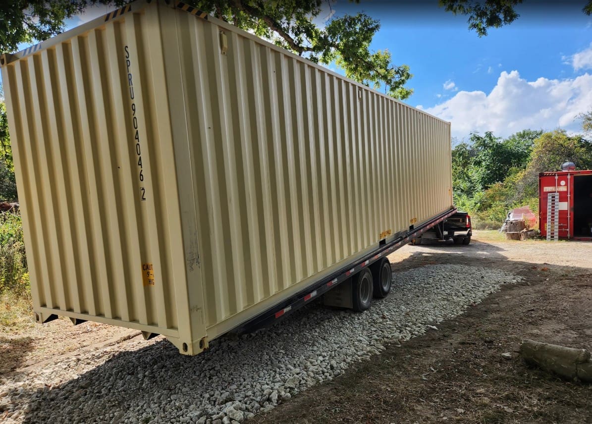 Tilt-bed delivery truck lowering a shipping container onto a gravel pad at a residential property